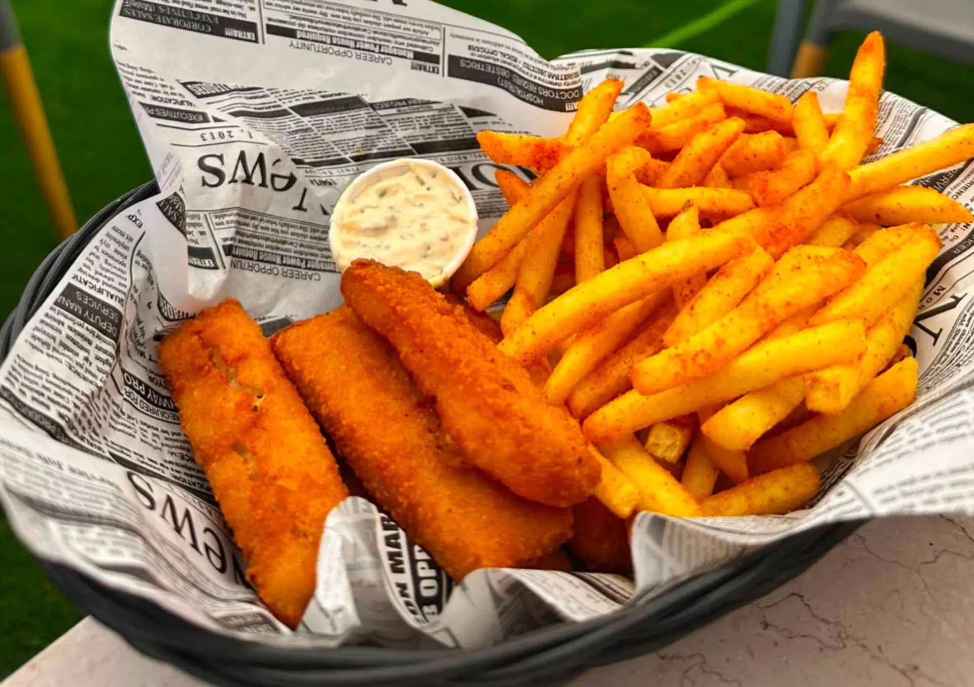 A newspaper-lined basket of breaded fish goujons with seasoned fries and tartare sauce at Zazoo