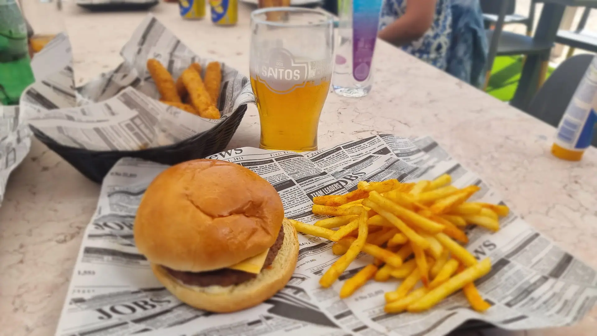 A cheeseburger with seasoned fries and a cold beer on a sunny Zazoo table