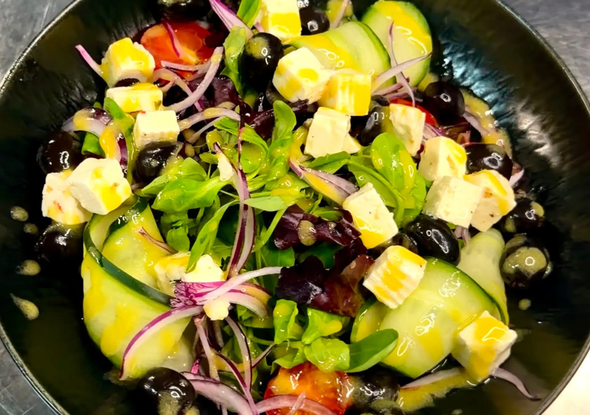 A fresh Greek-style salad with feta, olives and cucumber in a black bowl at Zazoo
